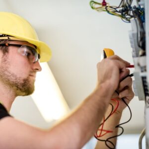 An electrician wearing a yellow hard hat and safety glasses using a circuit tester to check voltage levels in an electrical panel for safety and proper wiring.