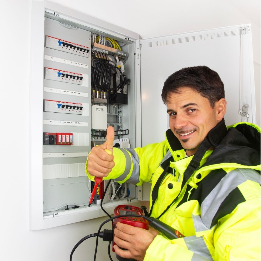 A commercial electrician wearing a high-visibility safety jacket gives a thumbs-up while inspecting an electrical panel, ensuring proper function and safety compliance.