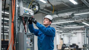 power installation: An electrician wearing a hard hat and safety glasses performing power installation work on a circuit breaker panel in a modern office setting.