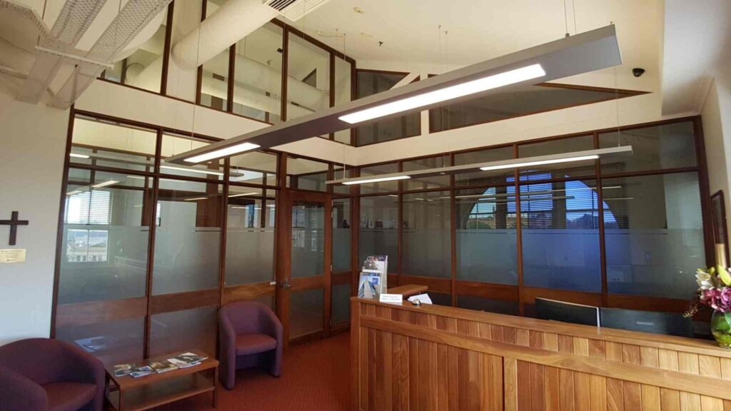 A professional office interior featuring modern suspended lighting fixtures above a wooden reception desk and frosted glass office partitions. The complexity of this integrated lighting and power system underscores the need for a certified commercial electrician to prevent and quickly resolve any electrical problems without compromising the high-end design.