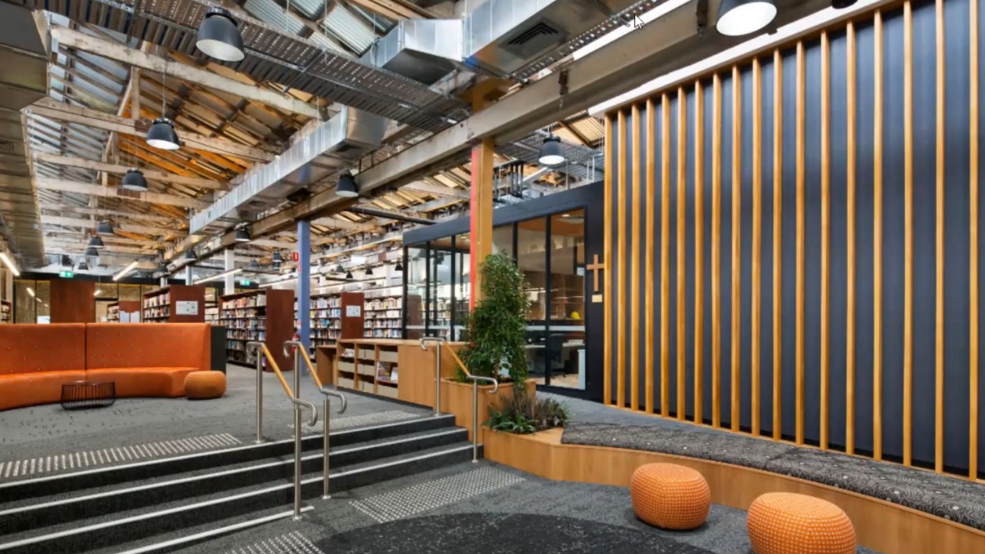 A spacious, multi-level commercial common area or library space with exposed industrial ceiling elements and various modern lighting fixtures. The area includes comfortable seating, shelves, and wood paneling, highlighting the need for specialized electrical maintenance to preserve both functionality and aesthetic appeal in a high-traffic Sydney commercial building.