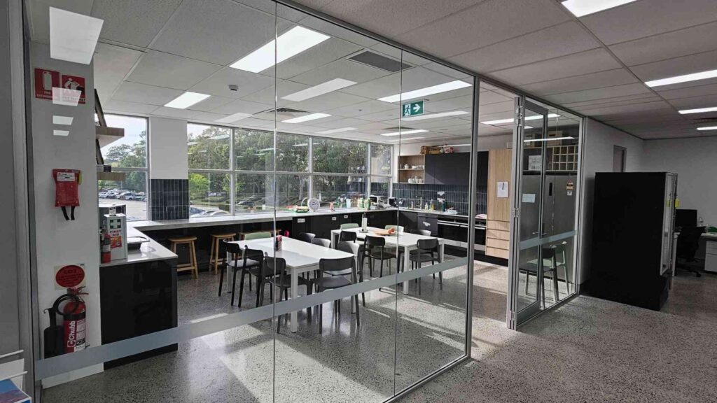 A modern Sydney office fitout featuring a bright communal kitchen and dining space with glass partitions and energy-efficient lighting. The layout promotes collaboration and a clean, open atmosphere.