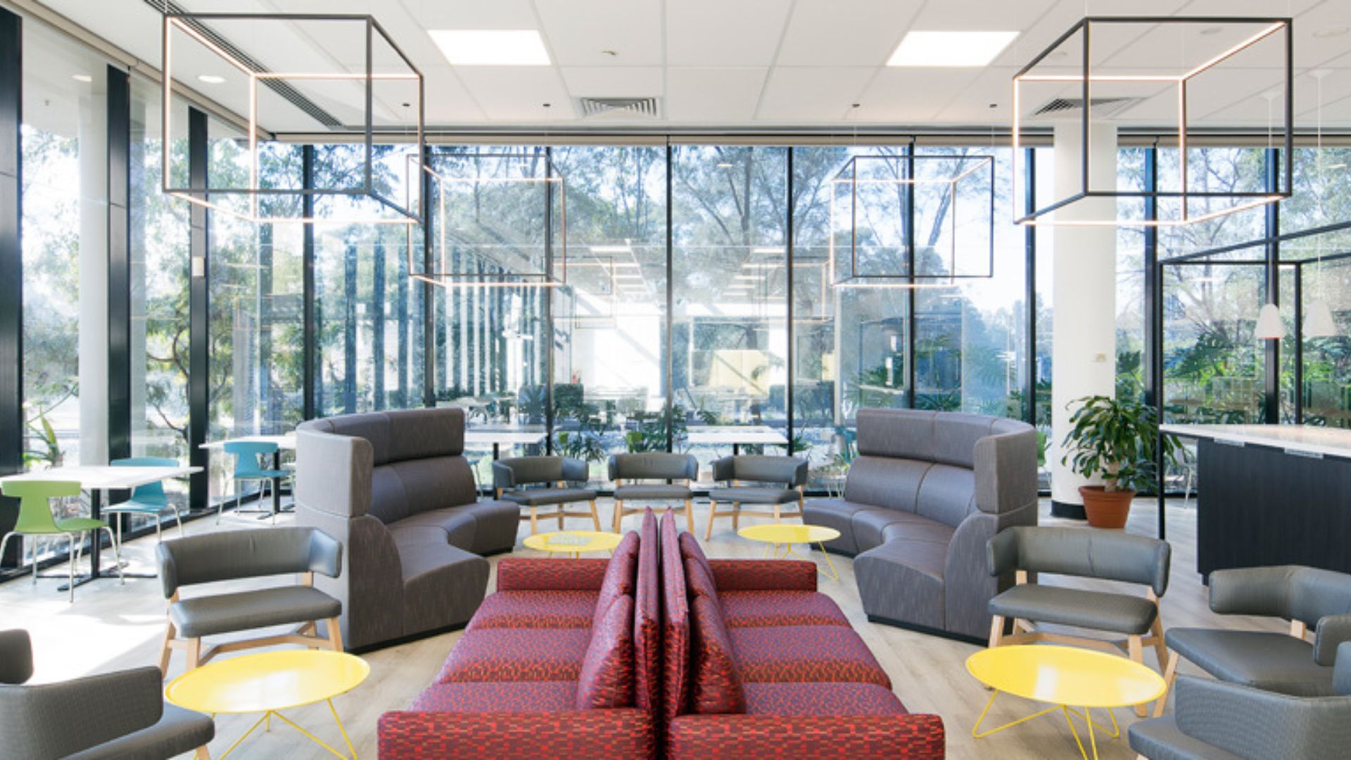 A modern, brightly lit office lounge with large floor-to-ceiling windows, featuring unique geometric cube pendant lights and contemporary furniture.